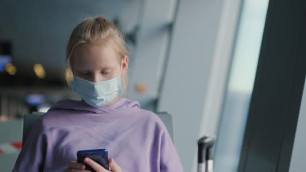 Дитина в захисній масці сидить в залі очікування аеропорту, використовуючи смартфон. Подорож під час пандемії коронавірусу — стокове відео
