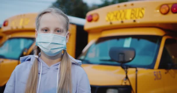 Porträtt av en skolflicka i en skyddande mask mot bakgrund av ett antal skolbussar — Stockvideo