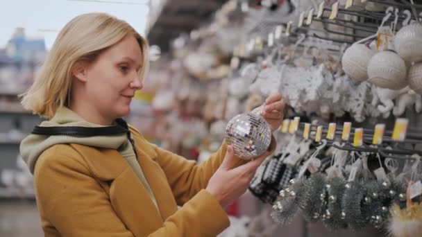 Uma mulher escolhe brinquedos para a árvore de Natal, segura uma bola brilhante em suas mãos. preparação para férias de inverno — Vídeo de Stock