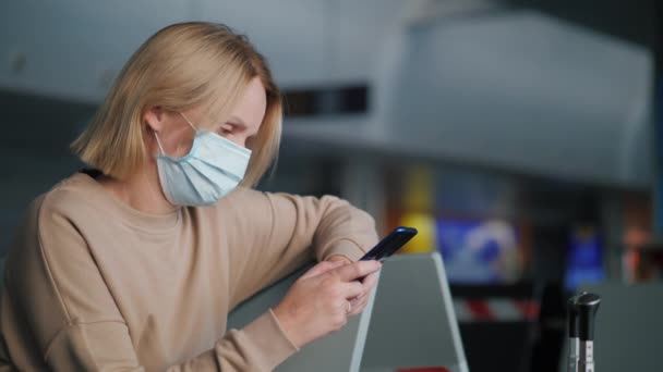 Een vrouw met een beschermend masker wacht op haar vlucht in de luchthaventerminal met een smartphone — Stockvideo