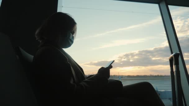 Silhouette d'une femme portant un masque de protection, assise à une grande fenêtre du terminal de l'aéroport, à l'aide d'un smartphone — Video