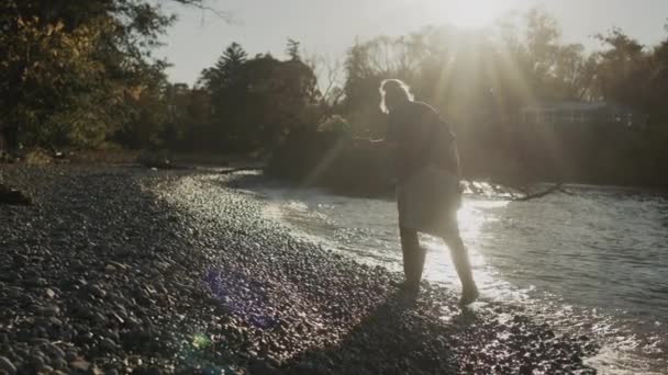 Egy gumicsizmás hajléktalan nő sétál a tó partján, keres valamit a parton.. — Stock videók
