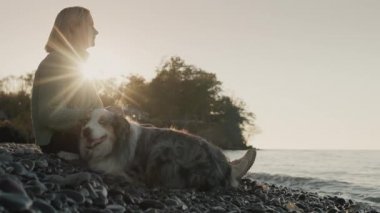 Sıcak kazaklı bir kadın gölün kıyısında bir köpeğin yanında oturuyor. Batan güneş onu aydınlatır ve sörf yapar.