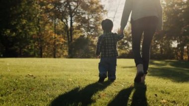 Anne, batan güneşin ışınlarında yeşil çimlerin yanında yürürken bebeği elinden tutar. Mutlu annelik ve çocukluk.