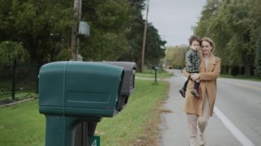 Kollarında çocuğu olan bir kadın posta kutusundan bir mektup alıyor.