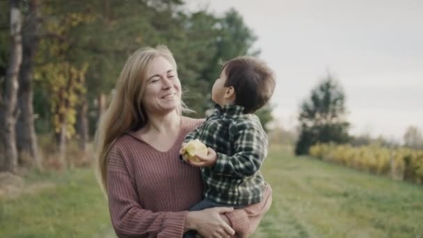Μια ευτυχισμένη μητέρα κουβαλάει ένα μωρό στην αγκαλιά της, τρώει ένα μήλο. Περπατούν στη φάρμα, τα σταφύλια φυτρώνουν κοντά. — Αρχείο Βίντεο