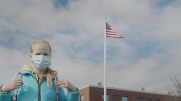 Шкільна дівчина в захисній масці стоїть навпроти шкільного та американського прапорів.. — стокове відео