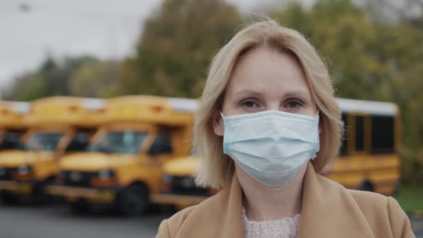 Portret van een lerares met een beschermend masker. Staat tegen de achtergrond van gele schoolbussen — Stockvideo