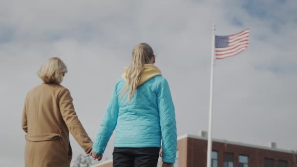 Mamá y su hijo se toman de la mano y miran la bandera estadounidense frente al edificio de oficinas — Vídeos de Stock