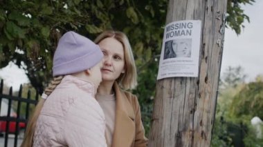 Çocuklu üzgün bir kadın, kayıp bir genç kadın ilanının verildiği direğin önünde duruyor. Kayıp kişileri ara
