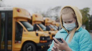 Koruyucu maskeli bir öğrenci sarı okul otobüslerinin yanında duruyor, akıllı telefon kullanıyor.