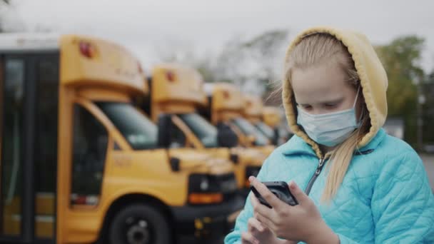 Uno scolaro con una maschera protettiva si trova vicino a una fila di scuolabus gialli, utilizza uno smartphone — Video Stock