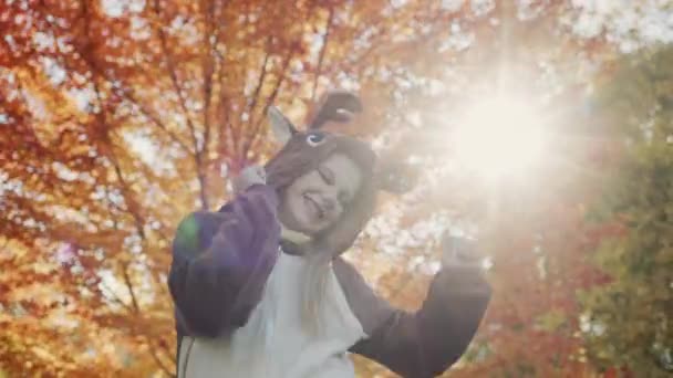 Ένα παιδί με στολή ελαφιού χορεύει ένα χαρούμενο χορό στο φόντο ενός όμορφου δέντρου με κίτρινα φύλλα. Ο ήλιος φωτίζει το κορίτσι από πίσω — Αρχείο Βίντεο
