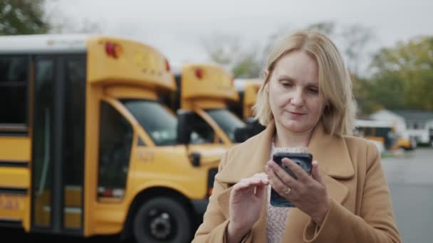 教師はスマートフォンを使い、黄色のスクールバスを背景に学校の庭に立つ. — ストック動画