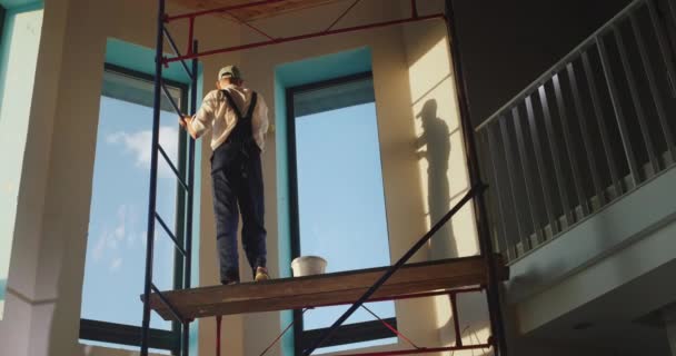 Будівельник в комбінезоні призначає стіну в будинку. Підставки на високому риштуванні — стокове відео