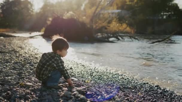 湖のほとりで遊んでいる小さなアジアの少年は、水に小石を投げます — ストック動画