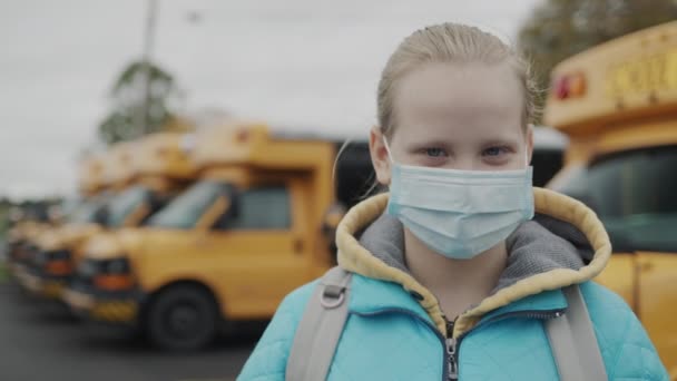 En skolflicka i en skyddande mask mot bakgrund av en rad gula bussar — Stockvideo
