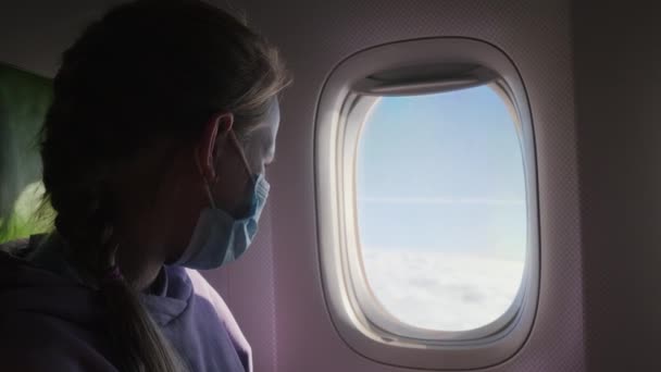Ребенок в защитной маске смотрит в окно самолета — стоковое видео