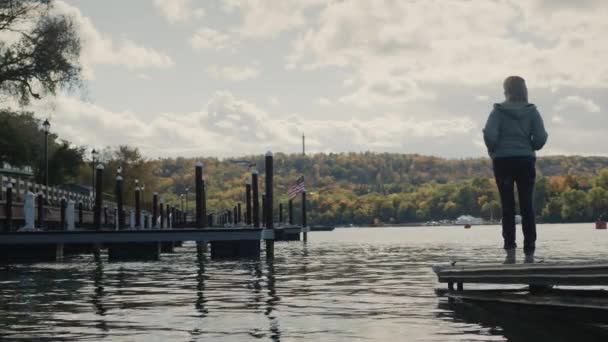 Uma mulher fica em um cais com vista para o Lago Ontário, a bandeira americana é visível à distância, não há outras pessoas e iates ao redor. Outono e fim da época turística. — Vídeo de Stock