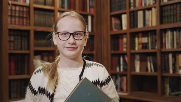 Porträtt av en 11-årig flicka med en lärobok, står mot bakgrund av bokhyllor i biblioteket — Stockvideo