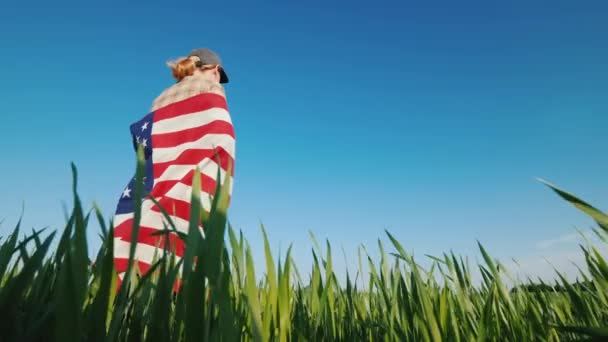 Uma mulher com uma bandeira dos EUA em seus ombros está em um pitoresco prado verde — Vídeo de Stock