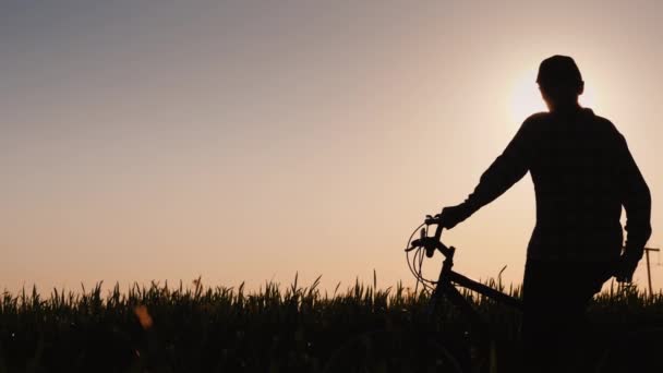 A silhueta de uma mulher com uma bicicleta de pé em um campo olhando para o pôr do sol — Vídeo de Stock
