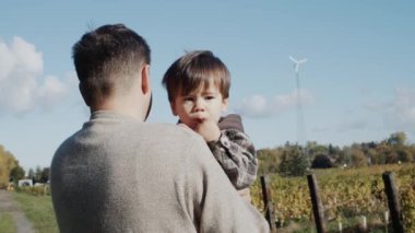 Kollarında küçük bir oğlu olan bir çiftçi bir üzüm bağının arka planında duruyor.