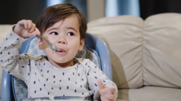 Cute Asian baby independently eats porridge from his plate, sits in a chair for feeding — Stock Video