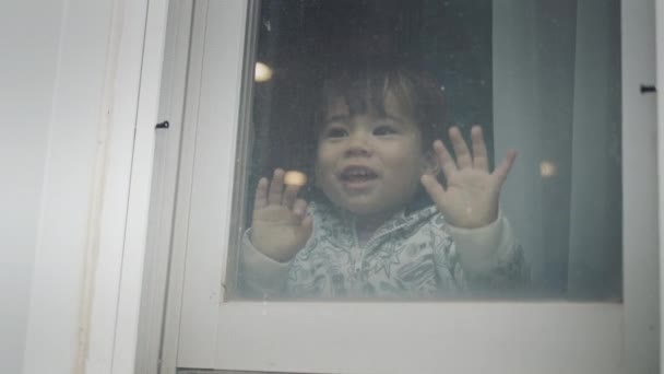 A happy two-year-old kid looks out the window, laughs happily — Stock Video