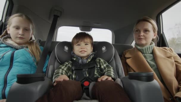 Una mujer con dos hijos conduce en el asiento trasero de un coche. Un niño en un asiento de coche para niños y una hermana mayor junto a ella — Vídeos de Stock