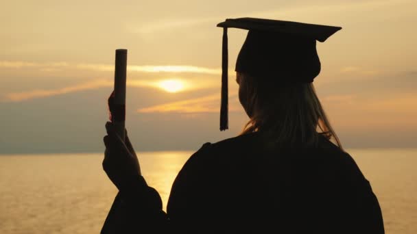 Een man met een diploma in zijn hand en een afgestudeerde kleding kijkt naar de zonsopgang boven de zee — Stockvideo