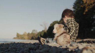 Çocuk gölün kıyısında oturur, suya çakıl taşları atar. Yanında küçük bir köpek yavrusu var..