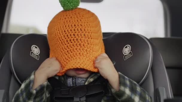 A cheerful kid plays hide and seek, pulls a hat over his head. Rides in a child car seat - a fun trip with a child — Vídeo de Stock