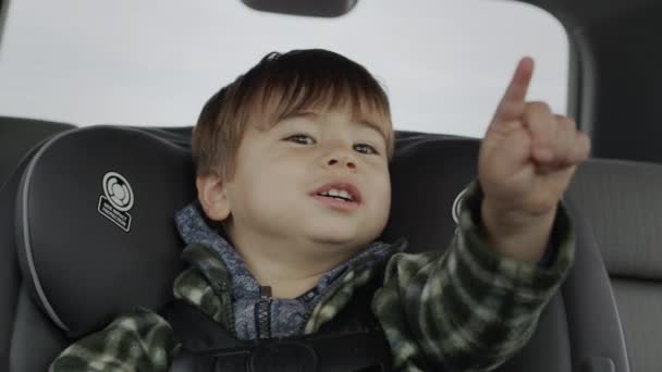 Een vrolijk kind van twee rijdt in een autostoeltje, wijst met zijn vingers naar voren. — Stockvideo