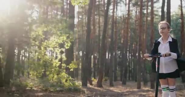 Skolflicka rider en skoter i en park med höga träd — Stockvideo