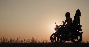 Genç bir çift gün batımına hayran bir şekilde motosiklette oturuyor.