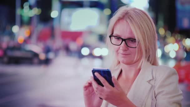 Young woman in glasses uses a smartphone on a busy street of Manhattan at night — Video