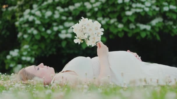 Cute pregnant woman lies on a lawn, holds a flower in her hand, dreams — Stock Video