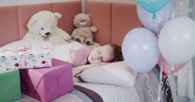 Bir çocuk yatağında, hediye kutuları ve balonların yanında uyur..