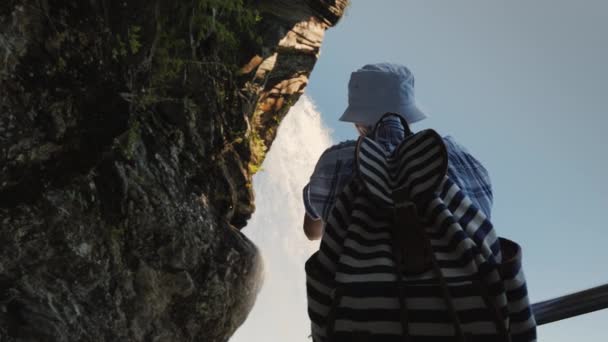 Steinsdalsfossen şelalesinin altında bir turist fotoğraf çekiyor, üzerinde bir kaya asılı ve bir su akıntısı akıyor. Düşük açılı çekim — Stok video