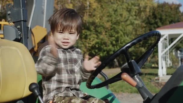 Pojken spelar entusiastiskt traktorförare. Sitter bakom ratten på en gammal traktor på gården — Stockvideo