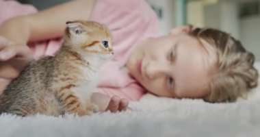 Çocuk bir kedi yavrusuyla oynuyor, evin zemininde battaniyeye uzanıyor.