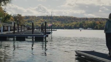 Ontario Gölü 'ne bakan bir kadın iskelede duruyor. Uzakta Amerikan bayrağı görülüyor. Etrafta başka kimse ve yatlar yok. Sonbahar ve turizm sezonunun sonu