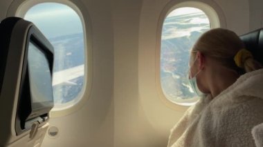 A child in a protective mask on his face looks out the window of the aircraft. Traveling during the covid 19 epidemic.