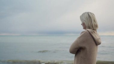 A woman in a warm knitted sweater looks at the sea where the storm begins.