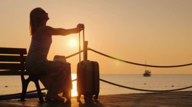 An adult woman enjoys relaxing on the beach. sits on a bench and clings to a suitcase on wheels.