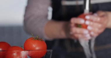 Bir kadın elinin arka planında bir kase domates musluktan akan suyun altında domates yıkıyor..