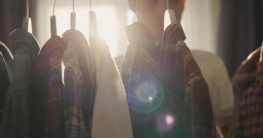 A young man chooses a shirt in a dressing room or shop. The sun from the window illuminates the rack with hangers