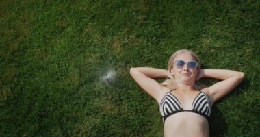 Attractive woman is escaping from the heat. Lies on the grass near the fountain of water from automatic watering. Slow motion video