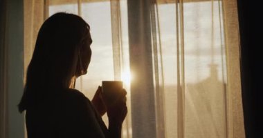 Elinde bir fincan çayla orta yaşlı bir kadın. Gün batımında pencereden dışarı bakıyorum..